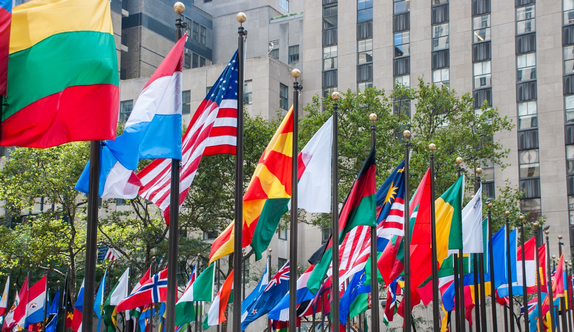 Colorful international flags displayed on poles in a city plaza with buildings in background.