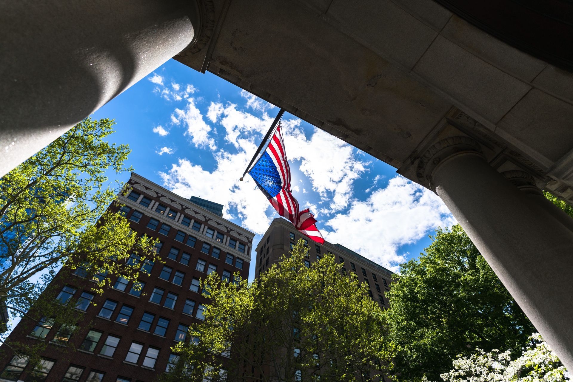 American flag waving beneath a bright blue sky with scattered white clouds, framed by stone columns