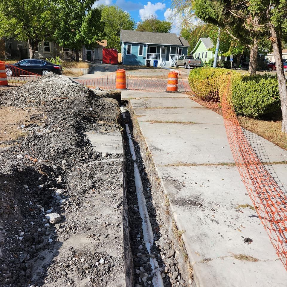 Construction site: trench dug alongside a sidewalk, orange barriers, houses in background.