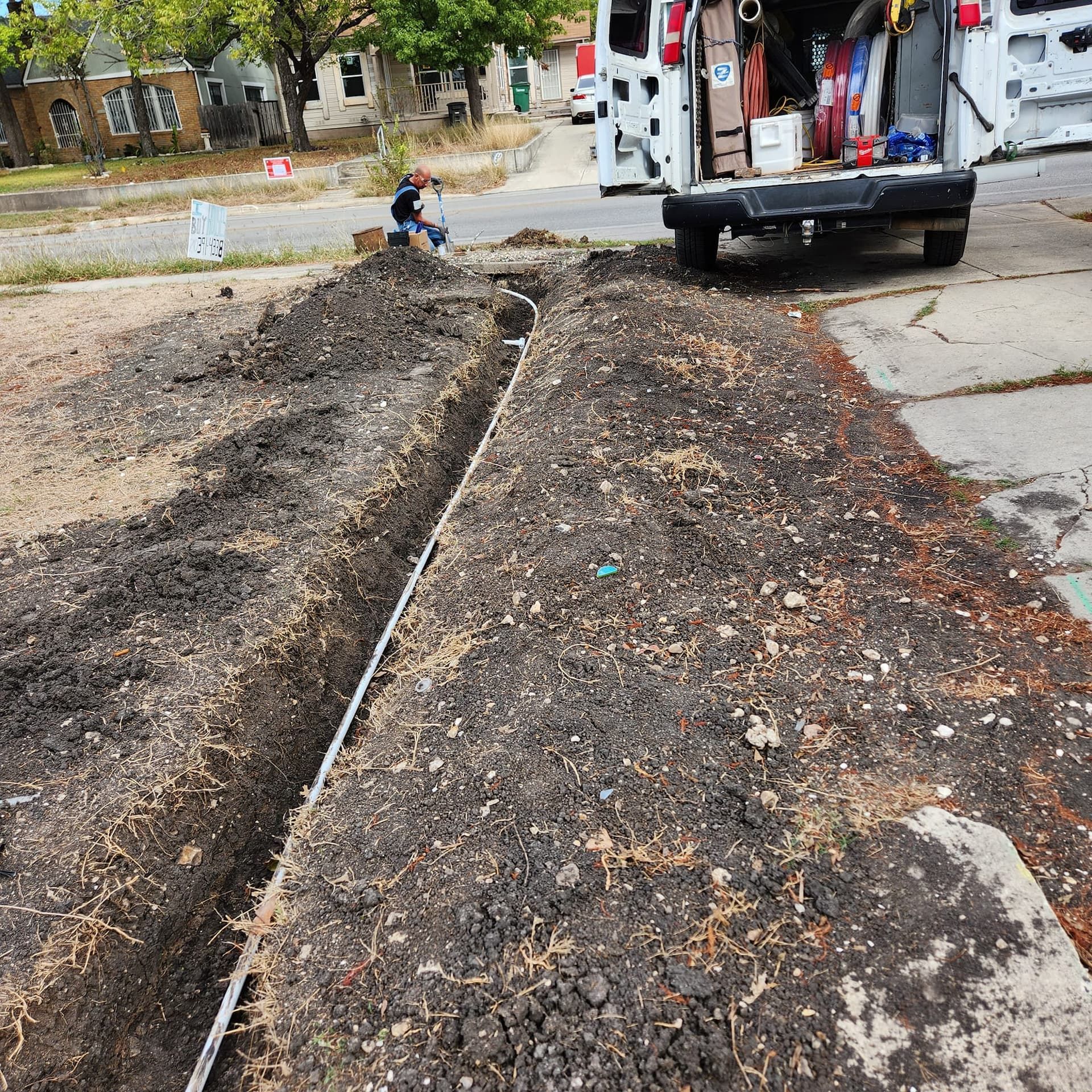 A trench dug in dirt, with a white pipe running through it. A van and a person are in the background.