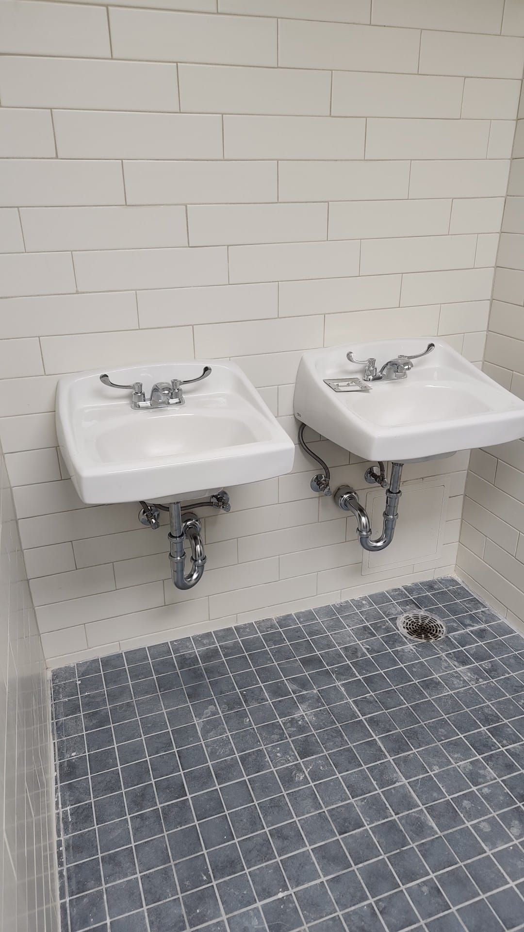 Two white sinks with chrome fixtures on a tiled wall, over a tiled floor with a drain.