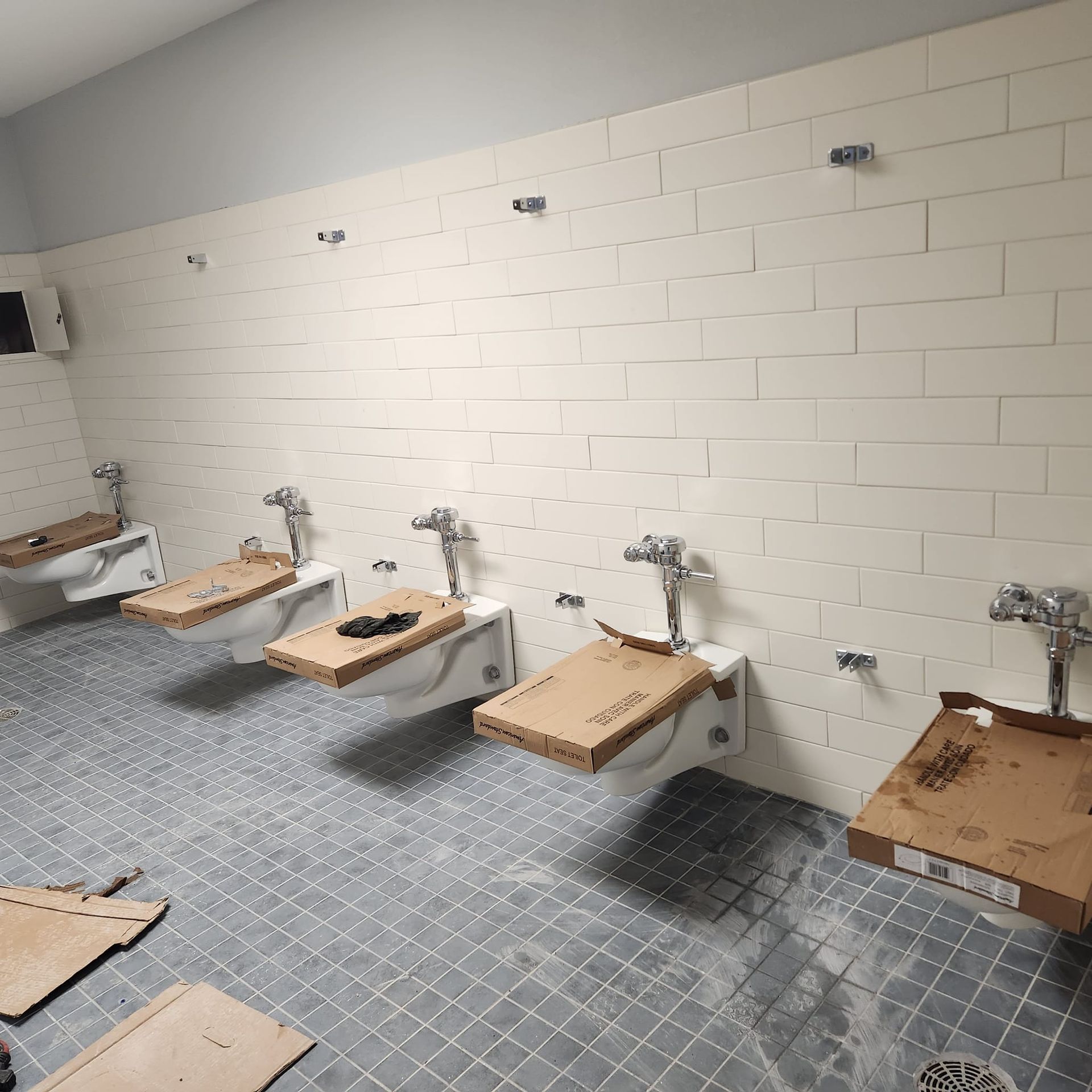 Public restroom under construction with white tiled walls, gray floors, and wall-mounted toilets.