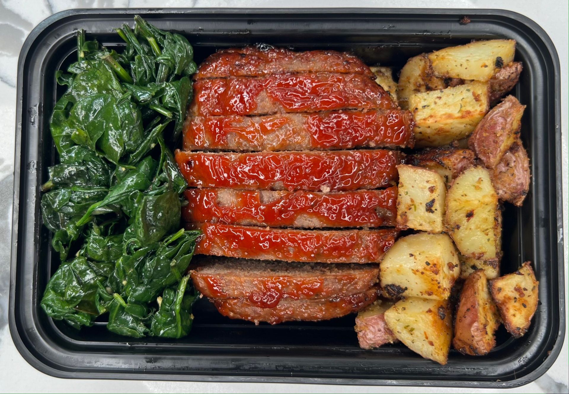 Meal prep container with sliced meatloaf, roasted potatoes, and spinach.