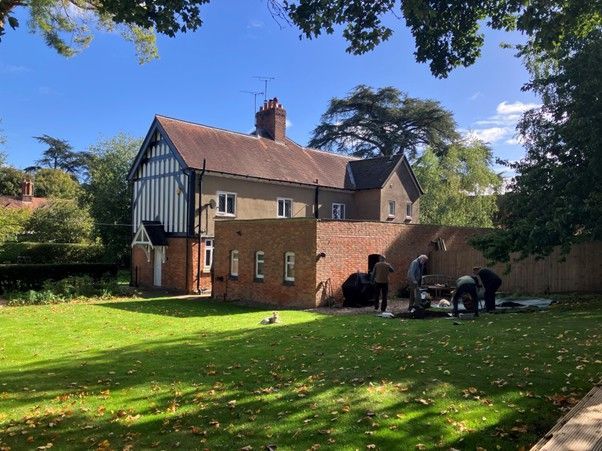 Green grass with a walled building and some people kneeling on the right hand side, the sky is blue and some tree branches are in the top of the image