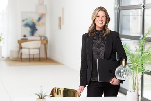 A woman in a suit is standing in front of a desk holding a laptop.