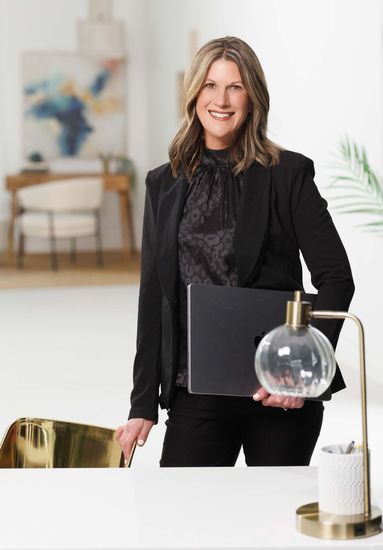 A woman in a black suit is standing in front of a desk holding a laptop.