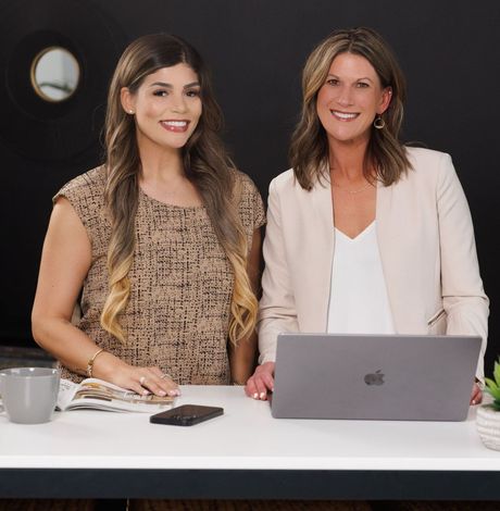 Two women are sitting at a desk with an apple laptop