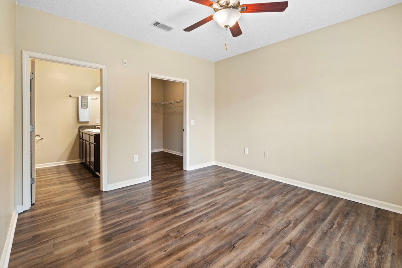 Empty bedroom with wood-look flooring, beige walls, a ceiling fan, and open closet and bathroom doors.