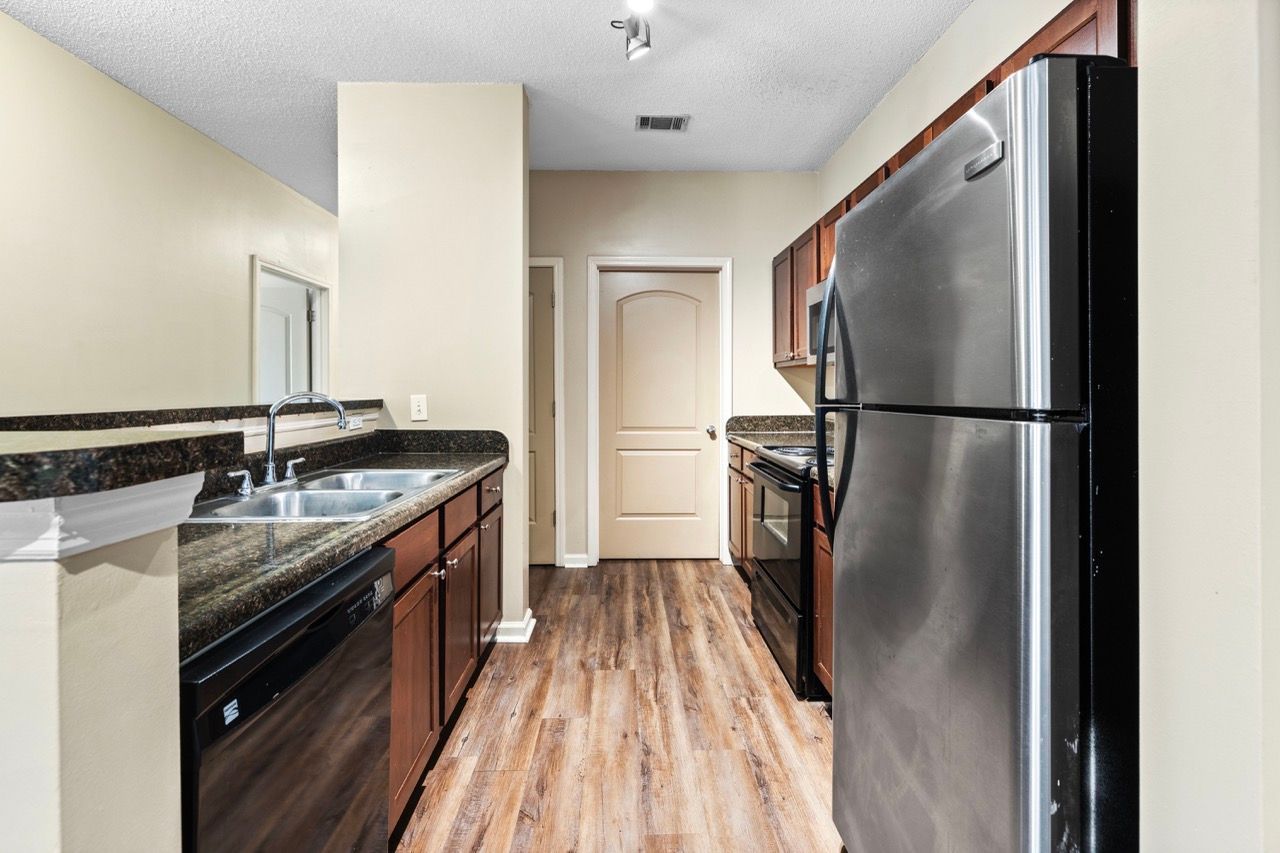 Galley-style apartment kitchen with stainless steel refrigerator, dishwasher, stove, and dark granite countertops.