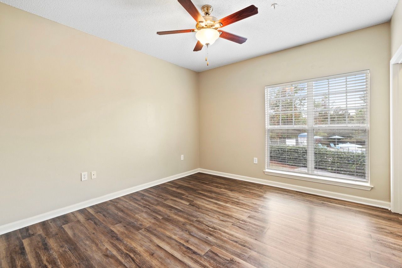 Empty living room with wood-look flooring, beige walls, a large window with blinds, and a ceiling fan.