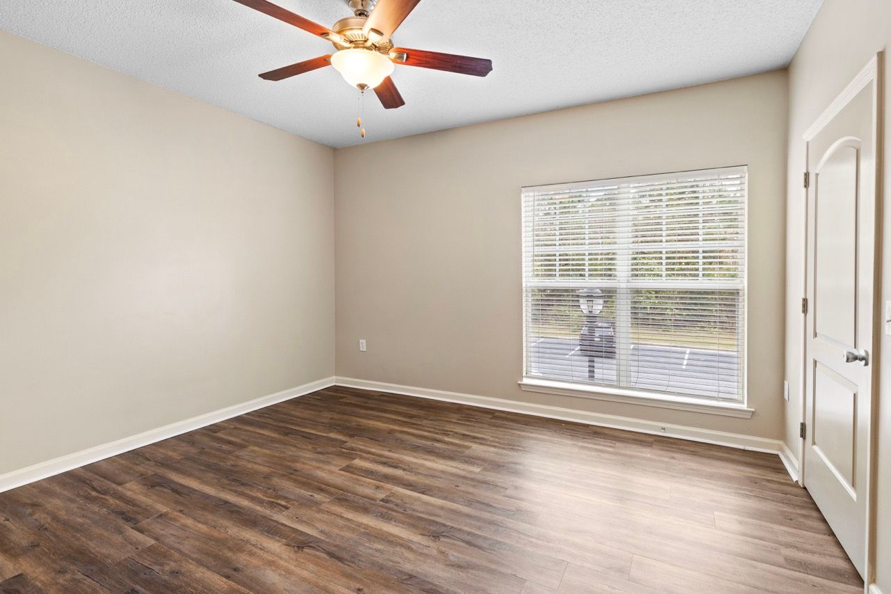 Empty living room with beige walls, wood-like laminate flooring, a large window with blinds, and a ceiling fan.