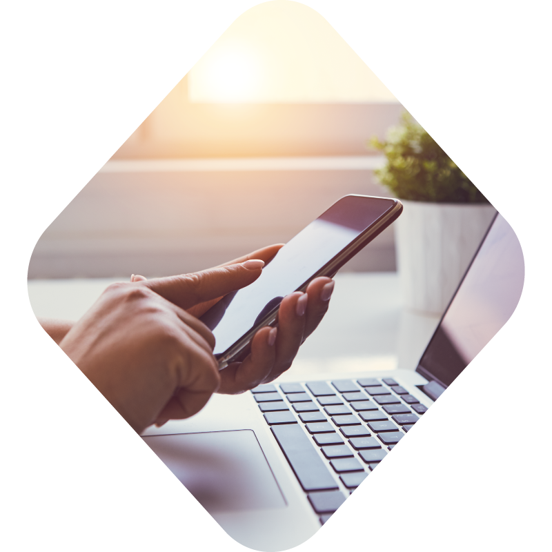 Photo of hands using a phone with a laptop also in view, all encapsulated in a diamond shape