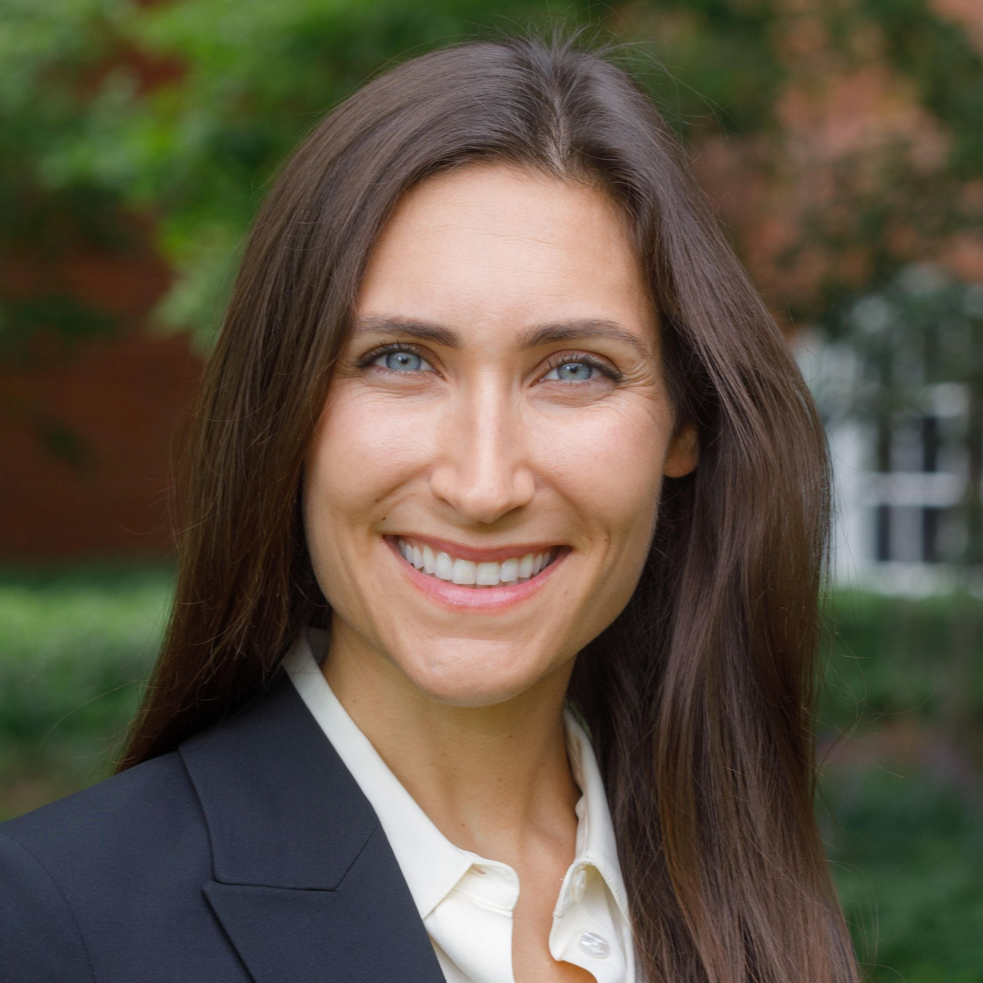 Woman in a blazer smiles at the camera outdoors, with long brown hair and blue eyes.