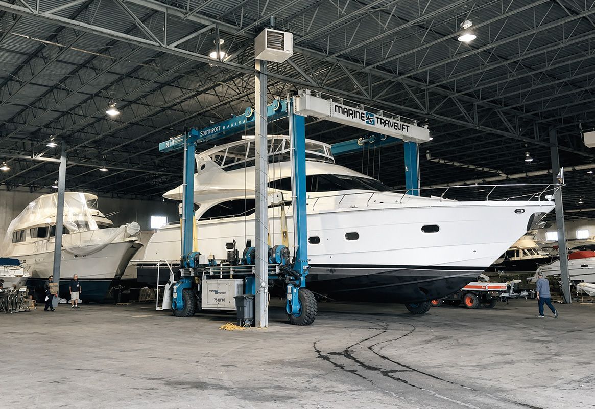 Yacht lifted in a boatyard by a blue travel lift, with another boat nearby under a covered hangar