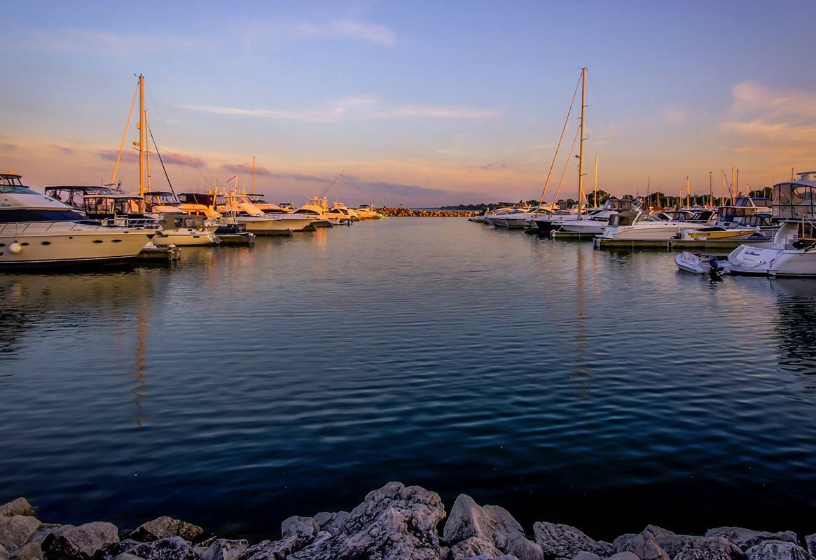 Marina at sunset with boats docked on calm water and a stone shore in the foreground