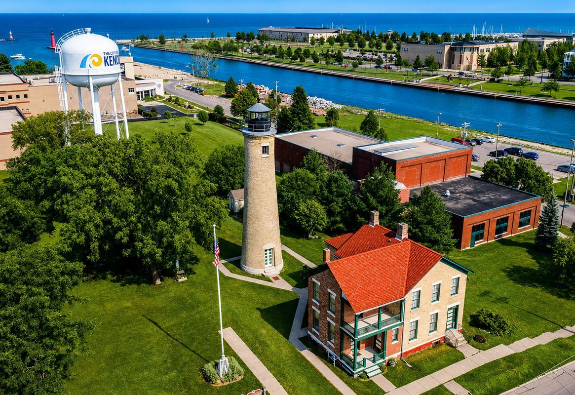Historic lighthouse and brick buildings beside a river with trees and a white water tower under a clear blue sky