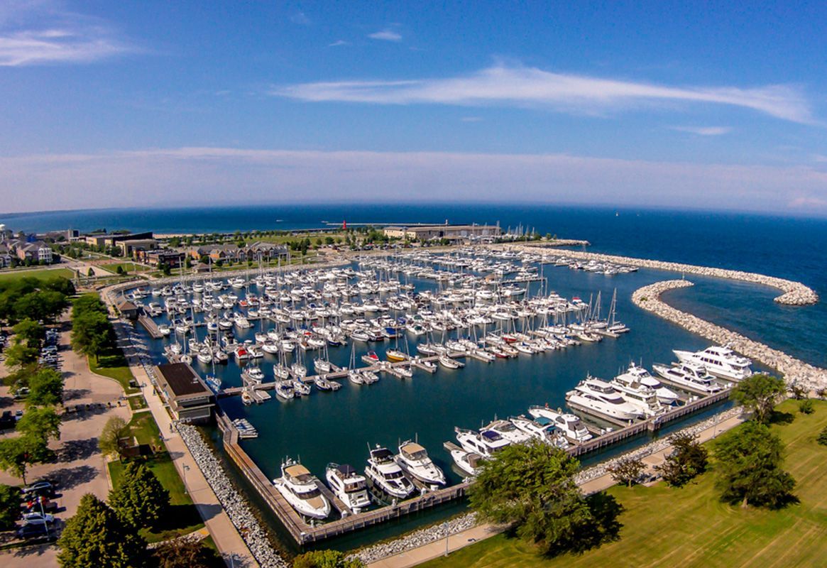 Aerial view of a marina packed with boats beside a curved breakwater and green shoreline under a blue sky