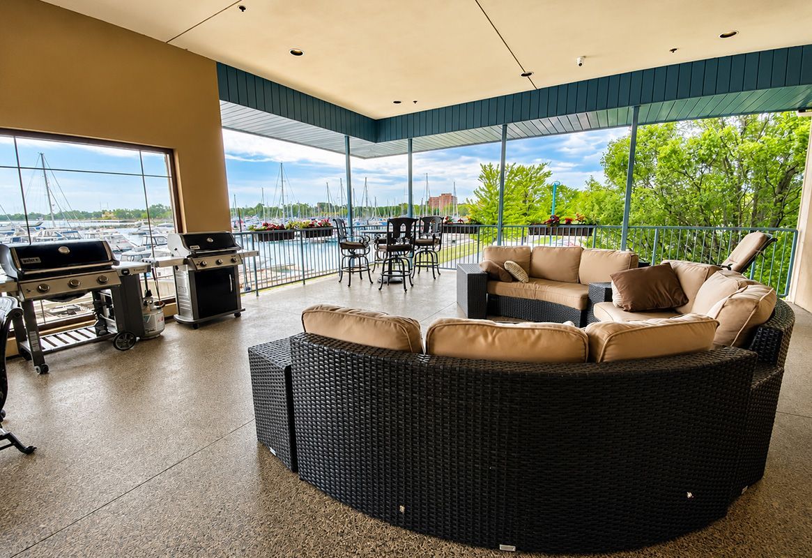 Covered outdoor patio with wicker seating, bar stools, and a grill overlooking a lake.