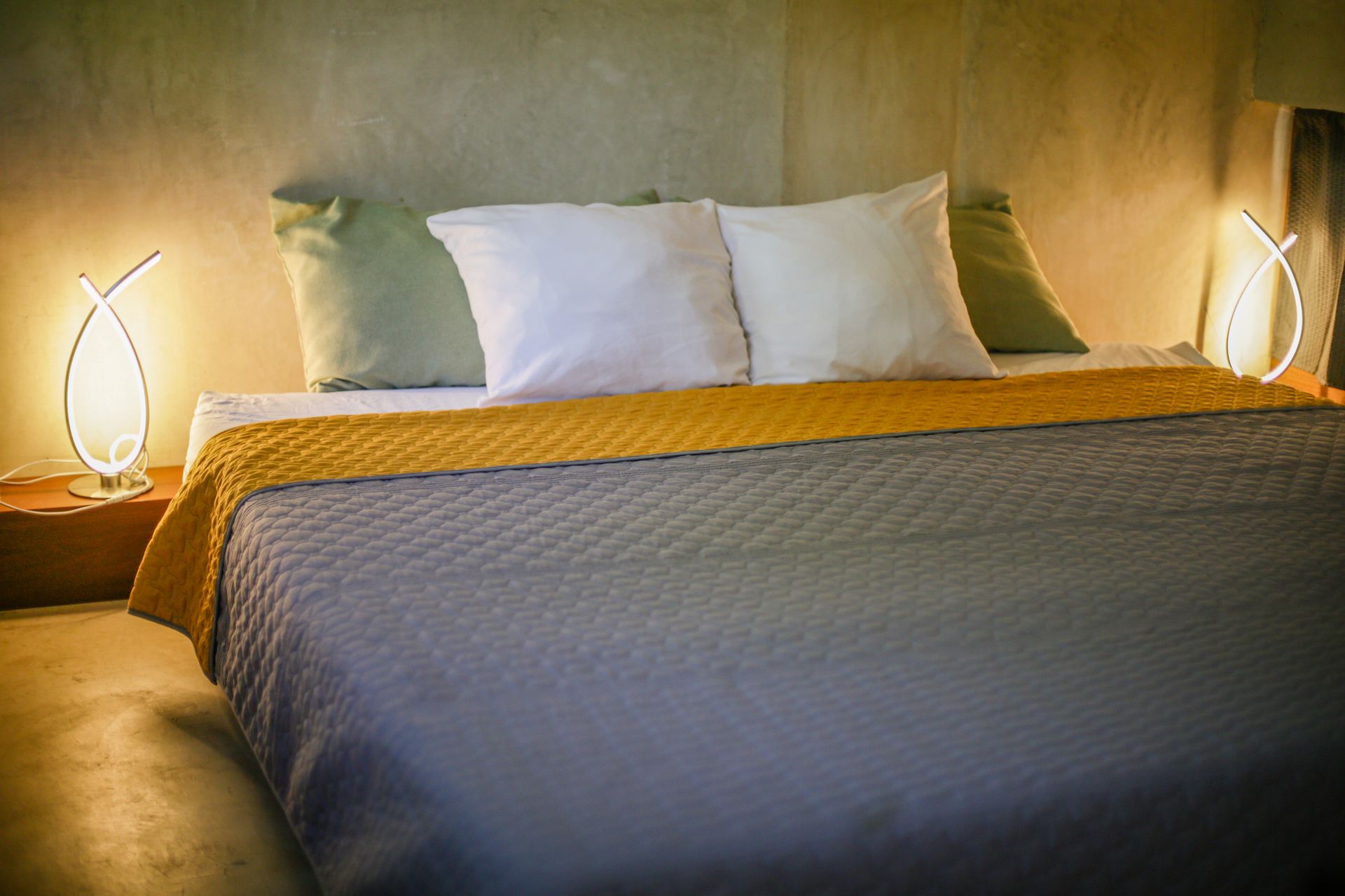 Cama con almohadas blancas y verdes, edredón gris, manta amarilla y dos lámparas curvas.