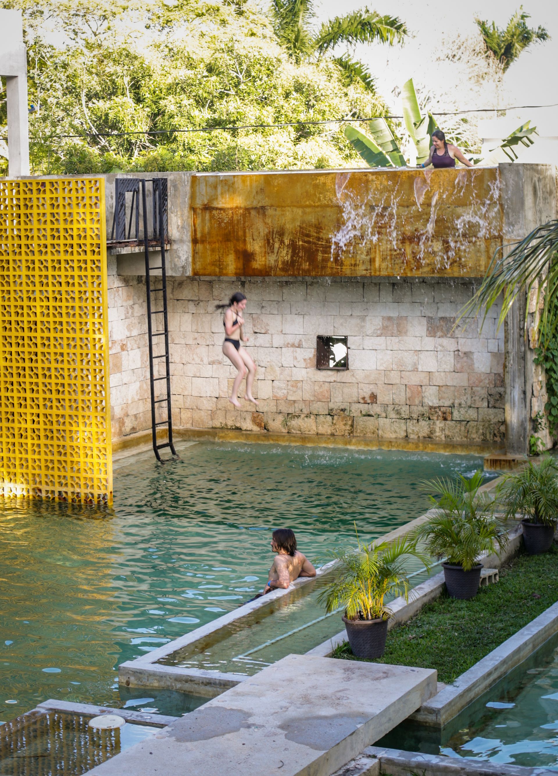 Mujer saltando a una piscina, otras personas en el agua y en el borde. Estructura amarilla, cascada, vegetación.