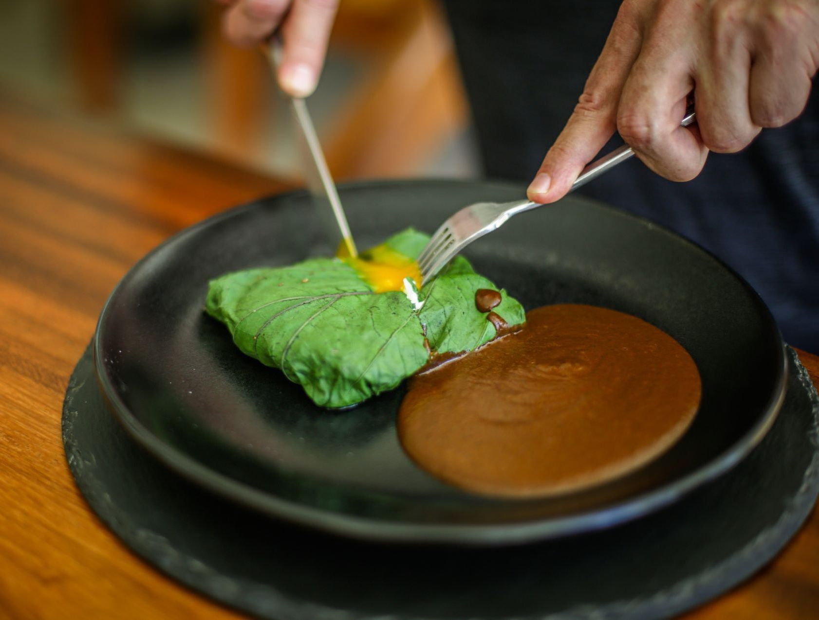 Persona cortando comida envuelta en una hoja verde, con una salsa marrón en un plato negro.