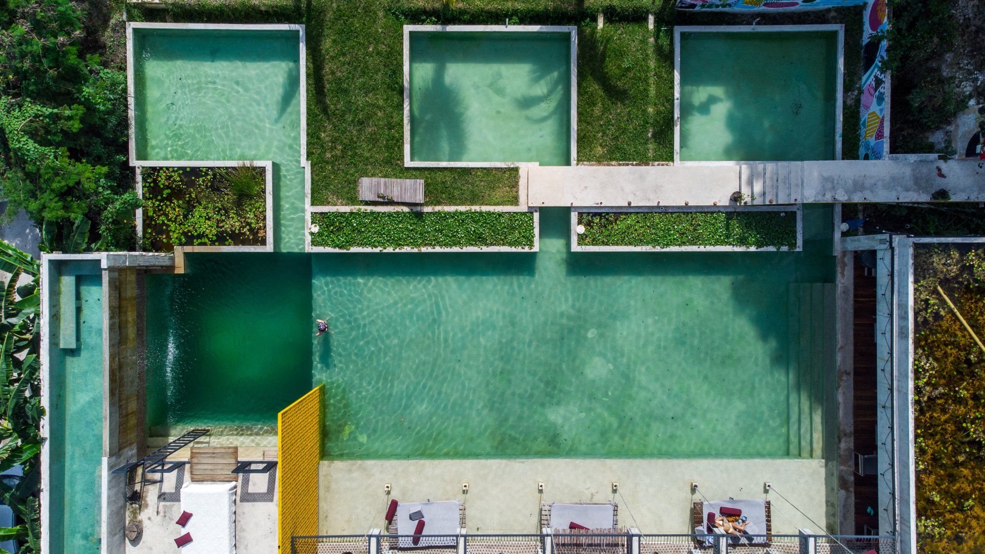 Vista aérea de una piscina del resort con agua turquesa, rodeada de camastros, vegetación y piscinas rectangulares de poca profundidad.