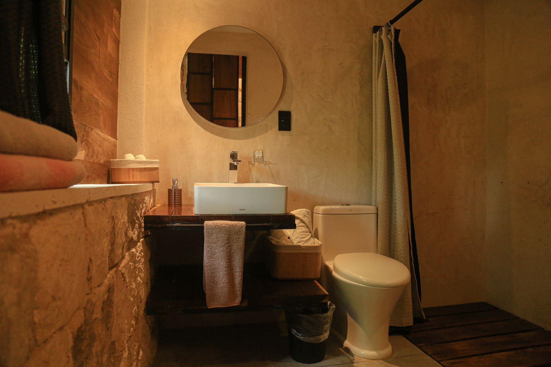 Baño con pared de piedra, espejo redondo, lavabo blanco y inodoro.