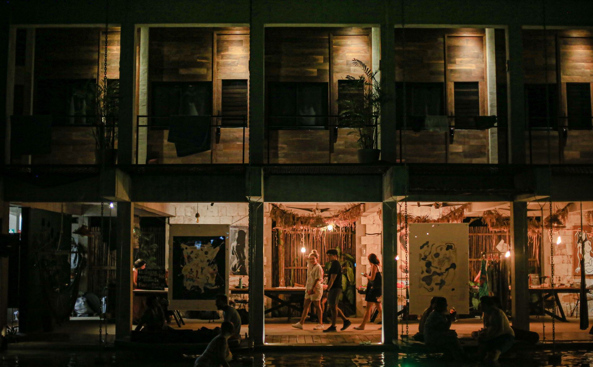 Exterior nocturno de un edificio con habitaciones abiertas; la gente se reúne en el interior, obras de arte en exhibición.
