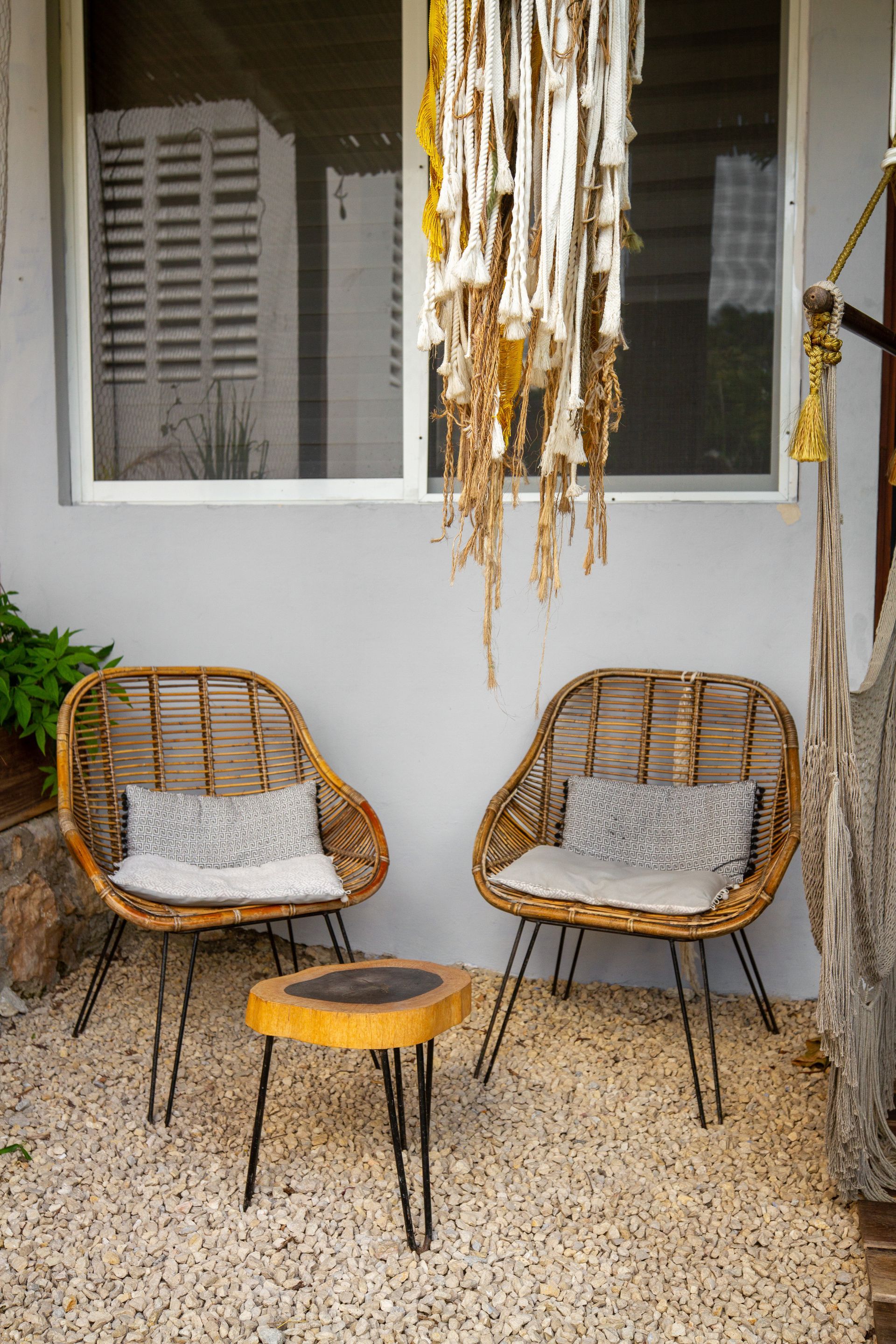Dos sillas de mimbre y una mesita de madera en un patio de piedra. Tapiz de macramé blanco y amarillo.