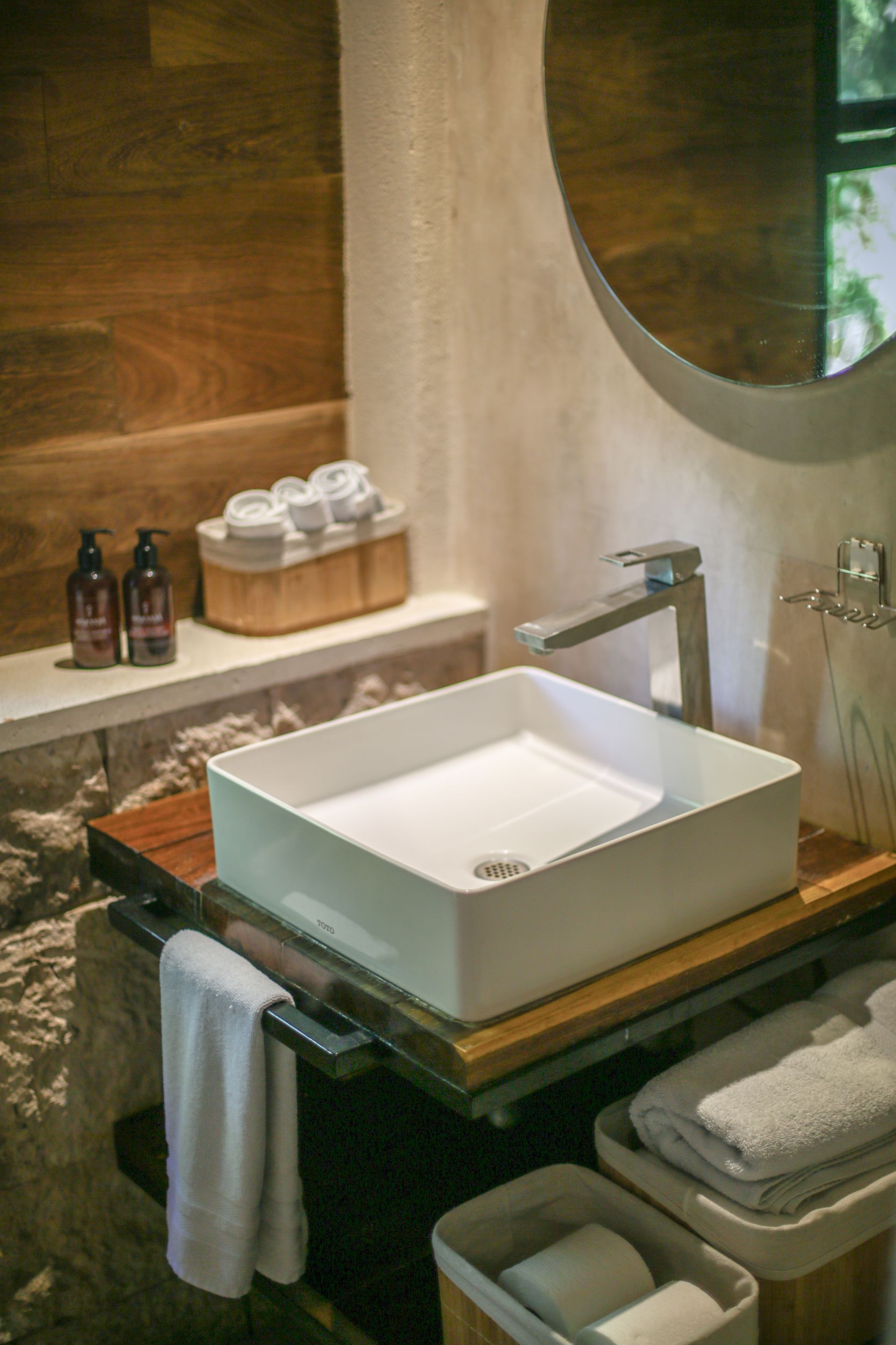 Baño moderno con lavabo cuadrado blanco, encimera de madera, pared de piedra, toallas y espejo redondo.