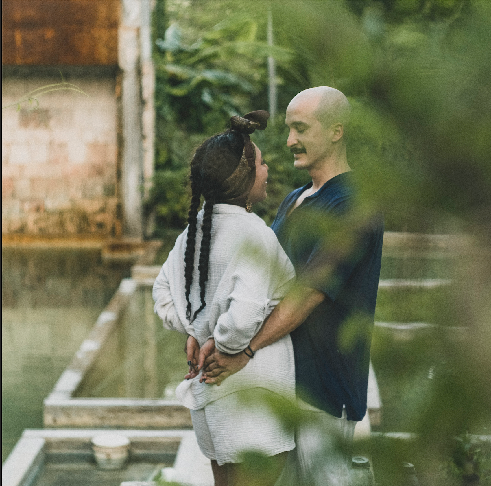 Pareja abrazándose al aire libre, mirándose con cariño. Mujer con trenzas y camisa blanca; hombre calvo y camisa azul. Vegetación, un entorno sereno.
