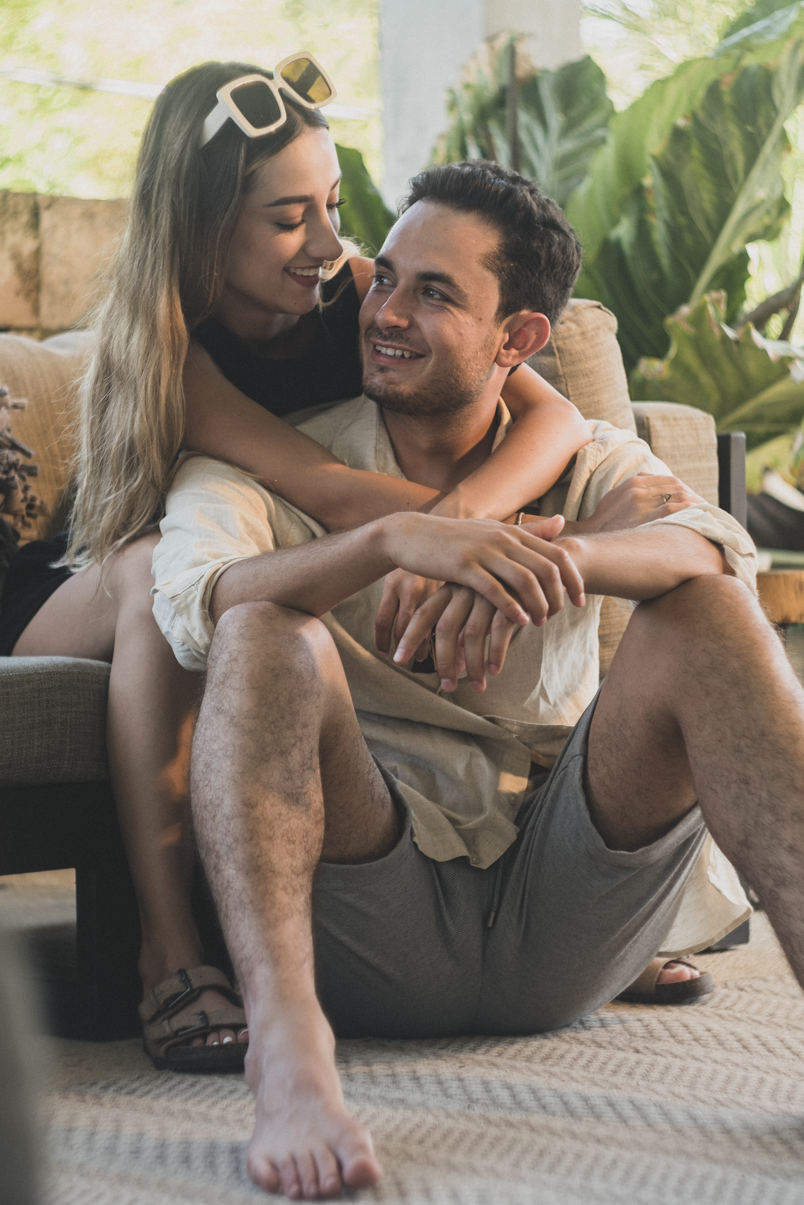 Una mujer abraza a un hombre, ambos sonriendo, en el suelo. Ambiente tropical, tonos beige y verdes.
