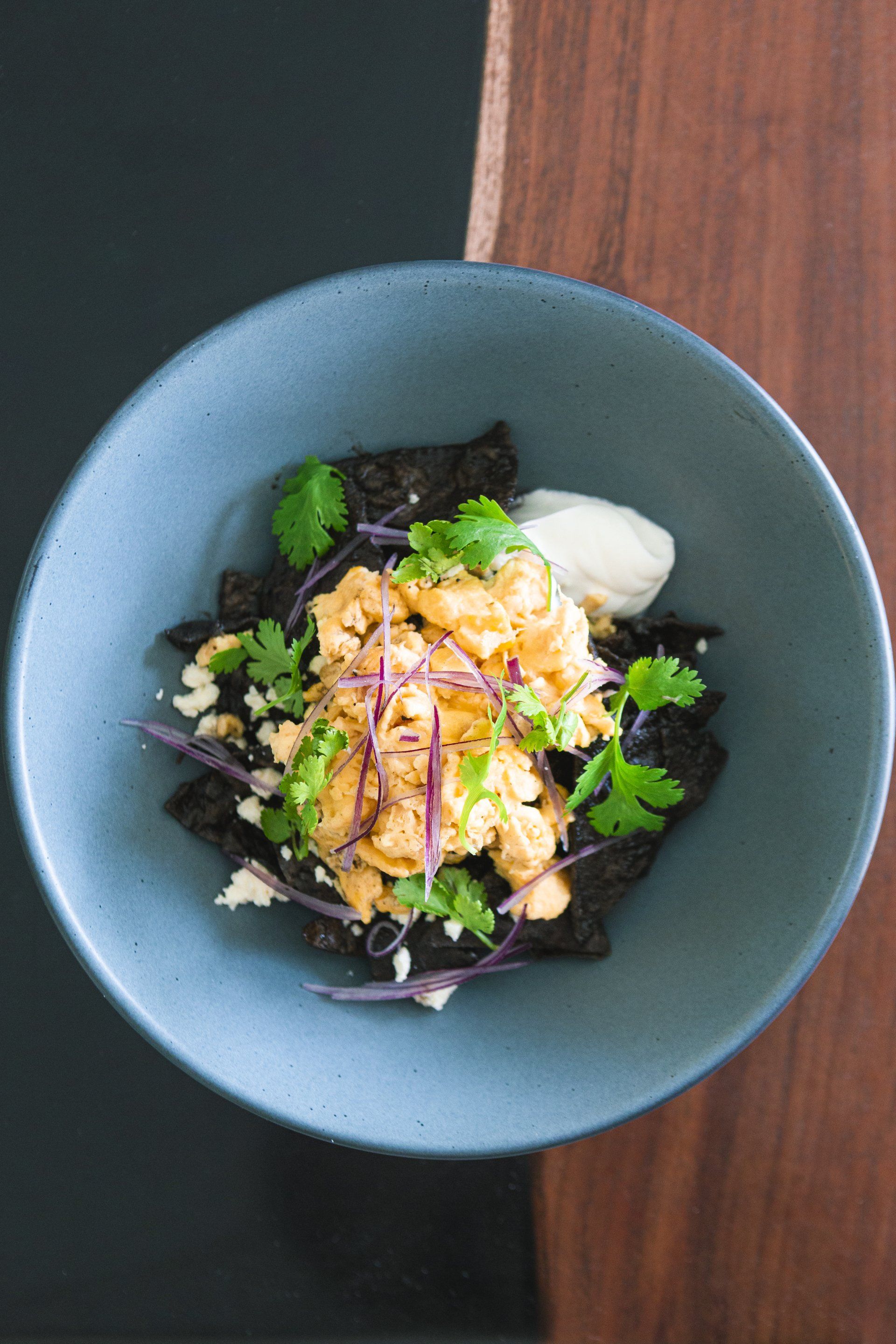 Tazón azul con huevos revueltos, patatas fritas negras, hierbas y una cucharada de salsa blanca.