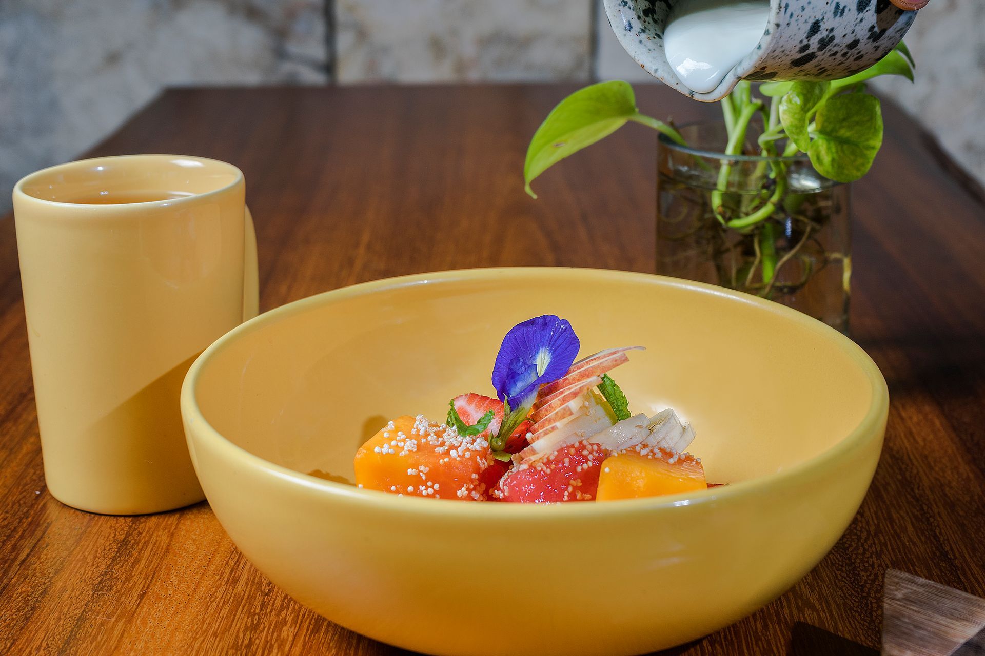 Cuenco amarillo de fruta con crema servida; taza amarilla, planta sobre mesa de madera.