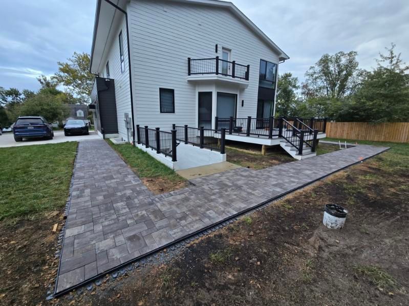 Two-story white house with brick pathways, black railings, and a wooden deck. Cars parked in the driveway.