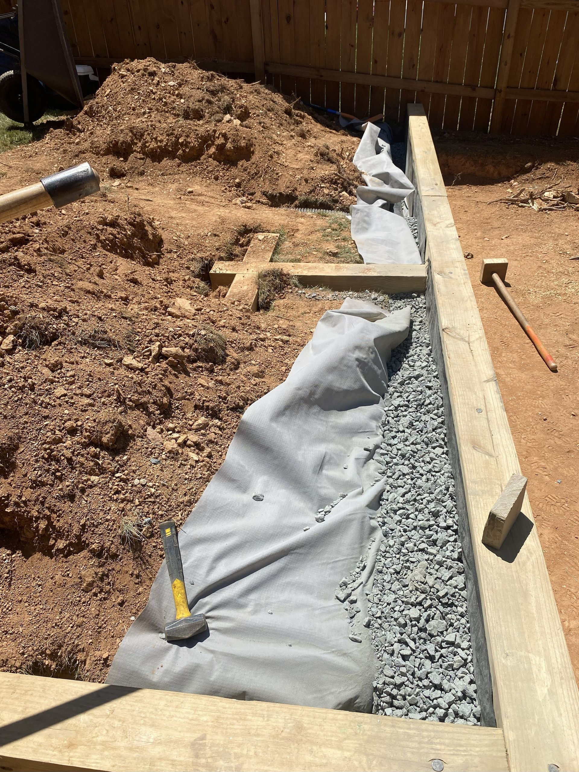 A gravel bed with landscape fabric, bordered by wooden beams, against a dirt pile and wooden fence.