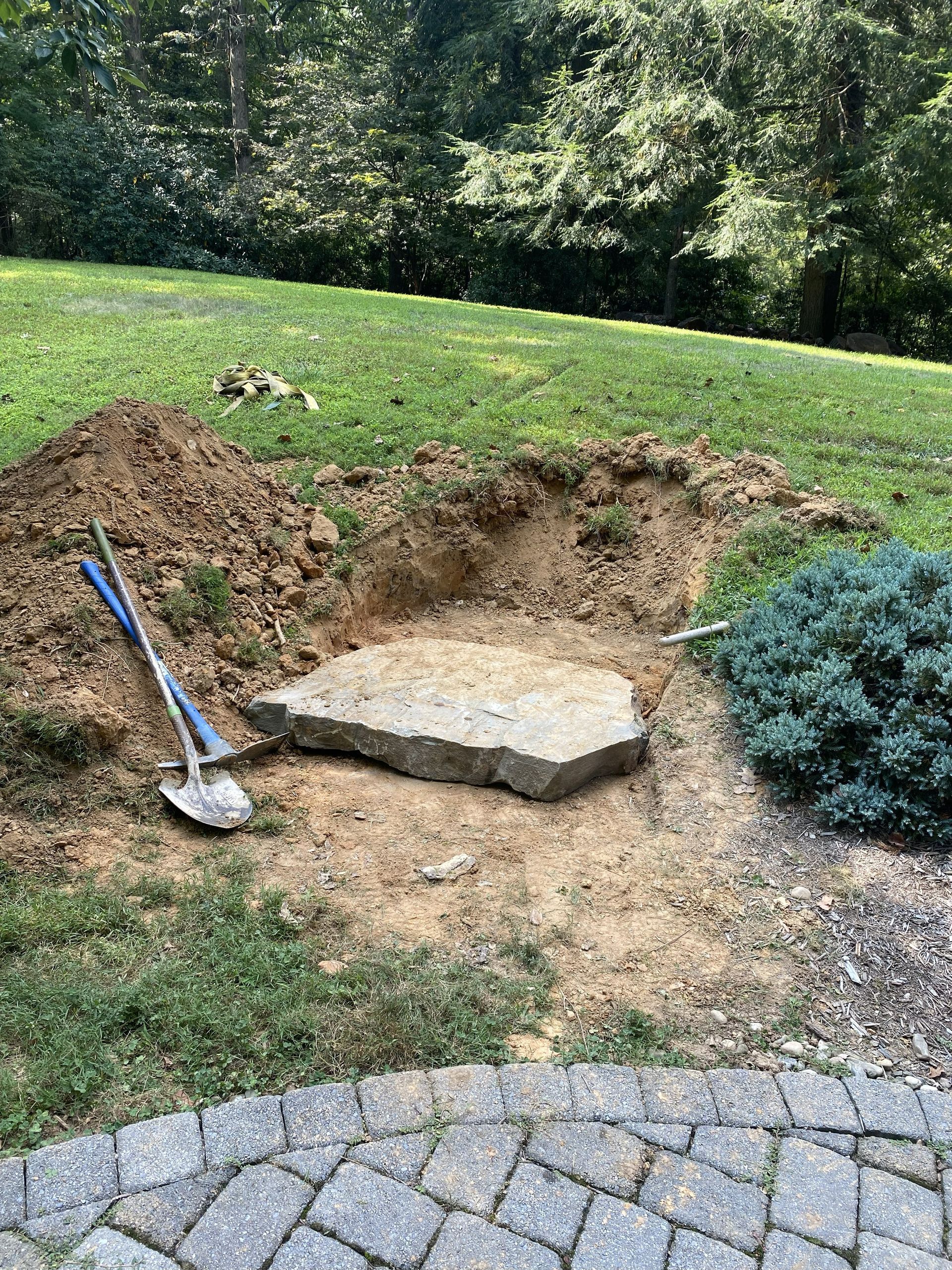 Hole dug in lawn with a stone slab and shovel. Ground is brown, grass is green, and the setting is outdoors.