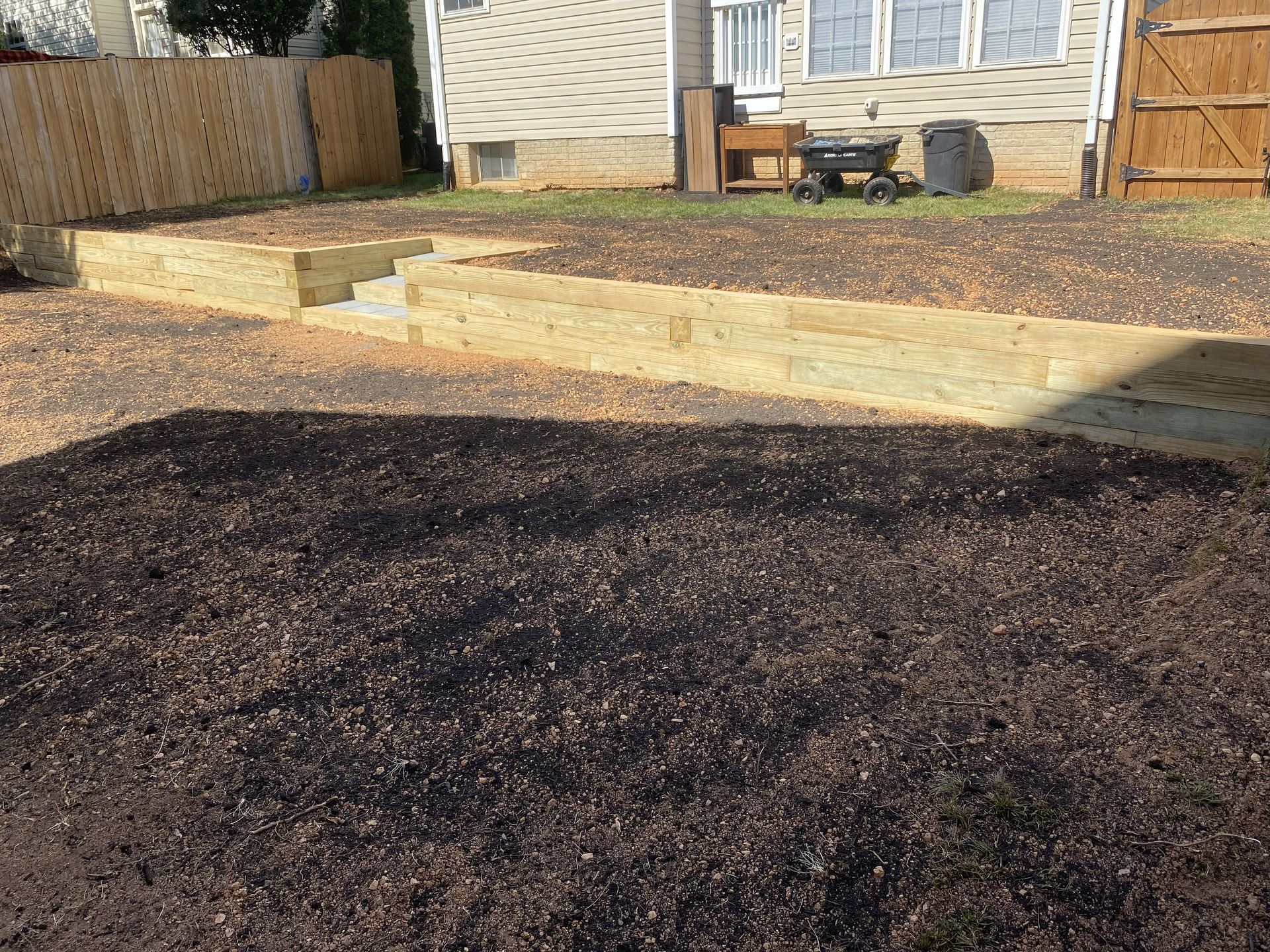 A backyard with tiered wooden retaining walls, mulch, and a house in the background.