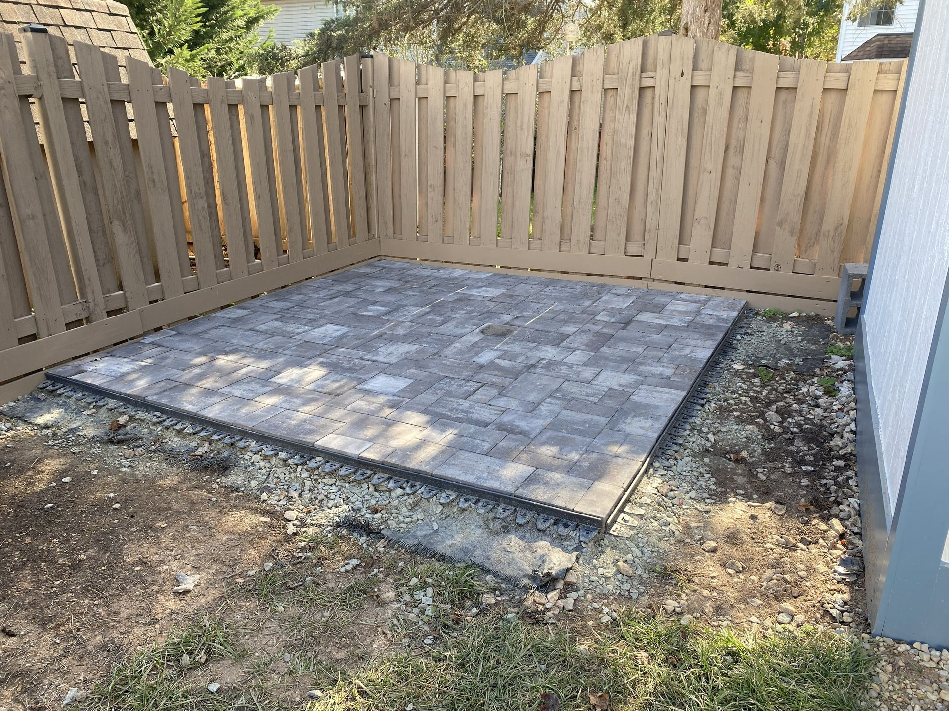 Paved patio area, bordered by gravel and a wooden fence; adjacent to a light-colored building wall.