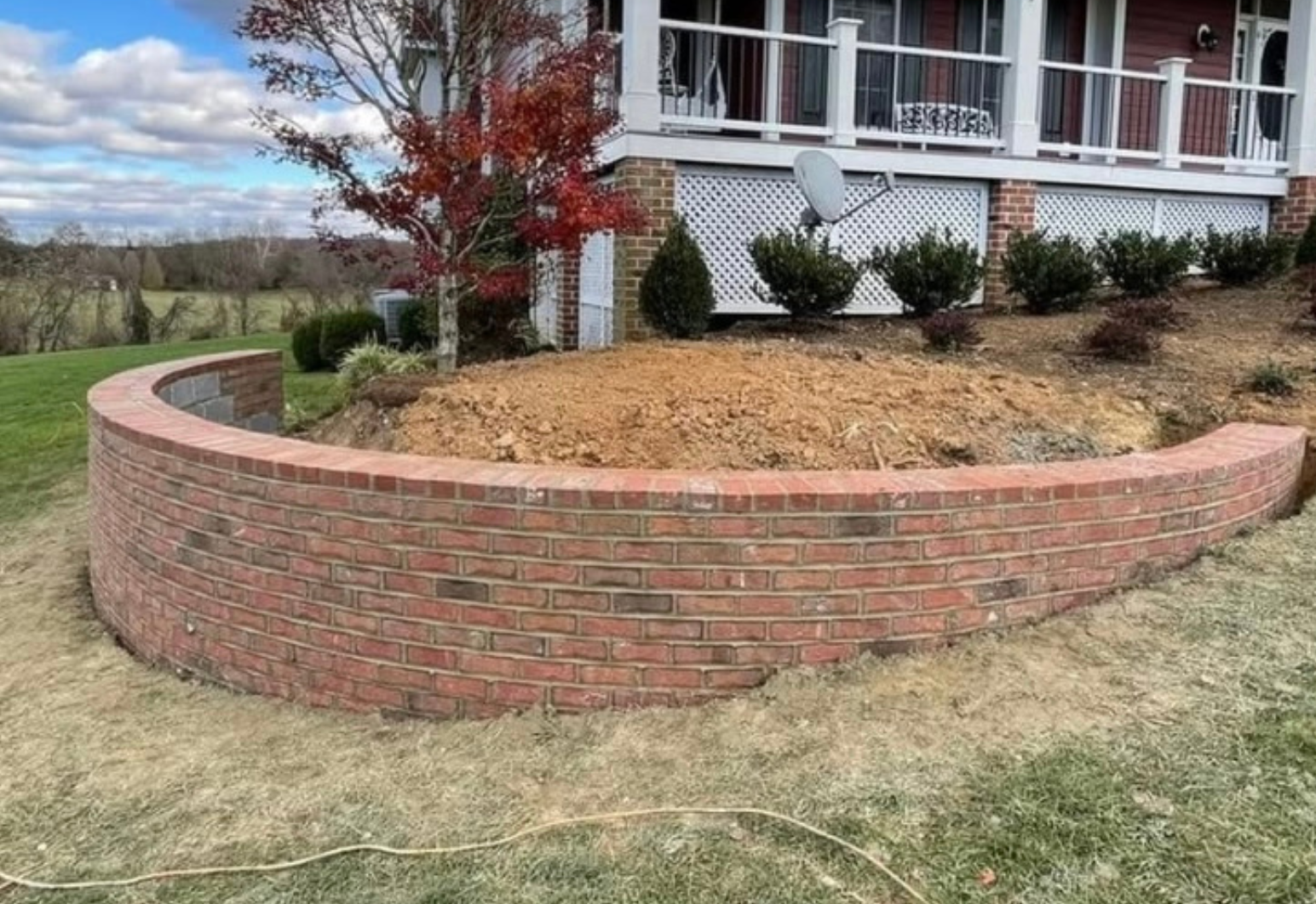 Curved brick planter with shrubs and mulch in front of a white house porch