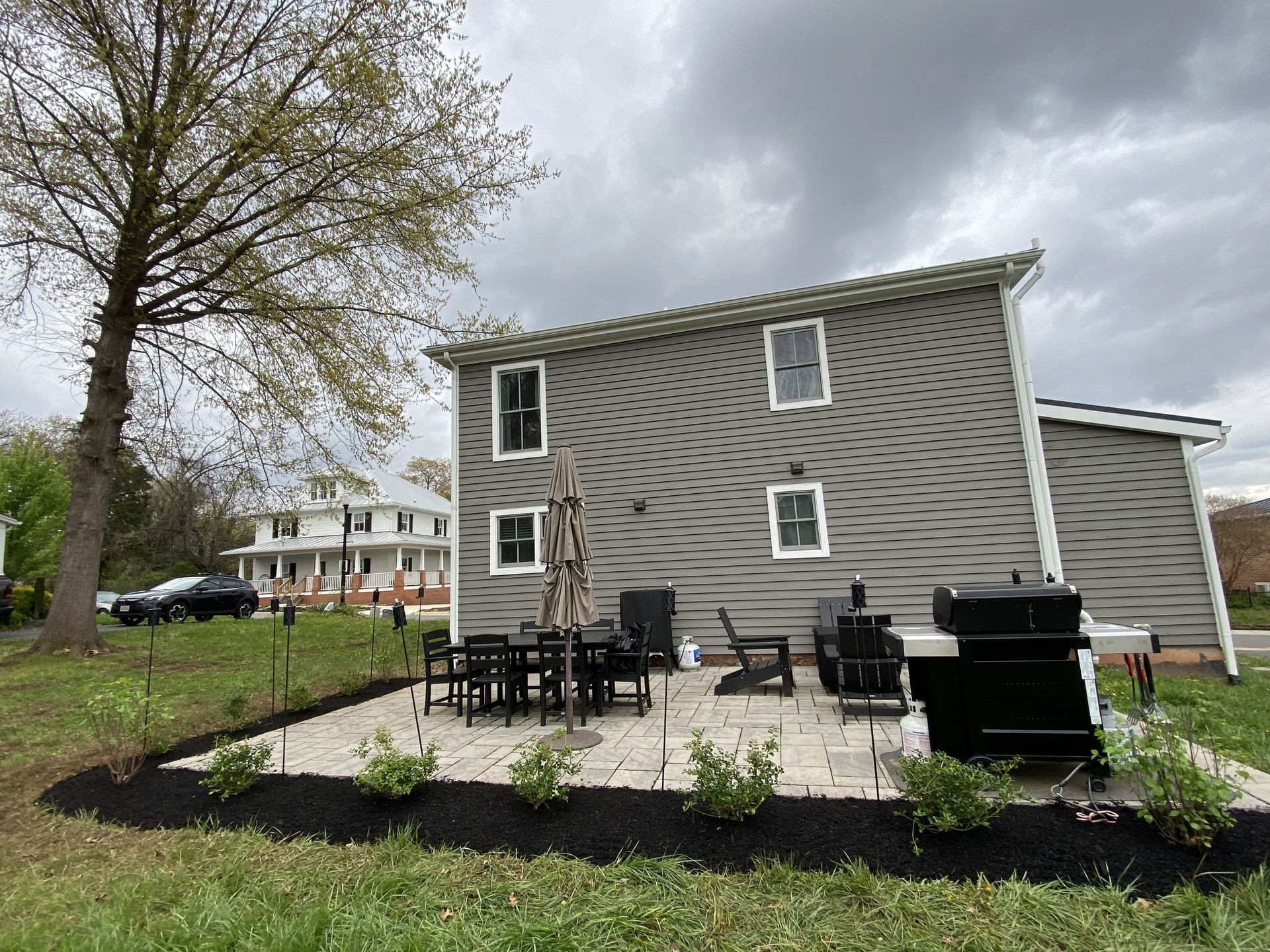 Backyard patio with gray house, table, grill, and dark mulch border under cloudy sky.