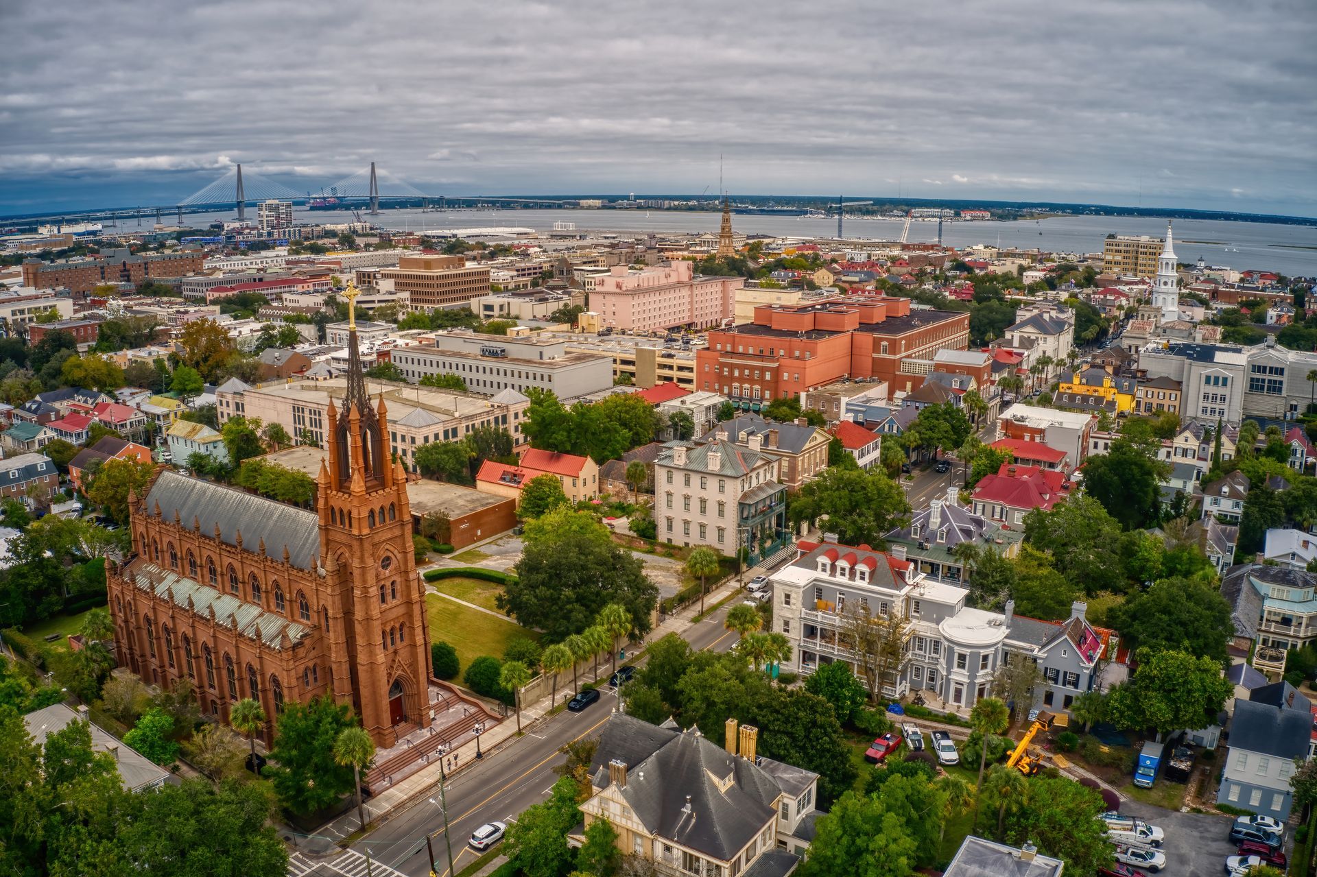 Aerial view of Charleston, SC, featuring a large red brick cathedral and waterfront cityscape under a cloudy sky.