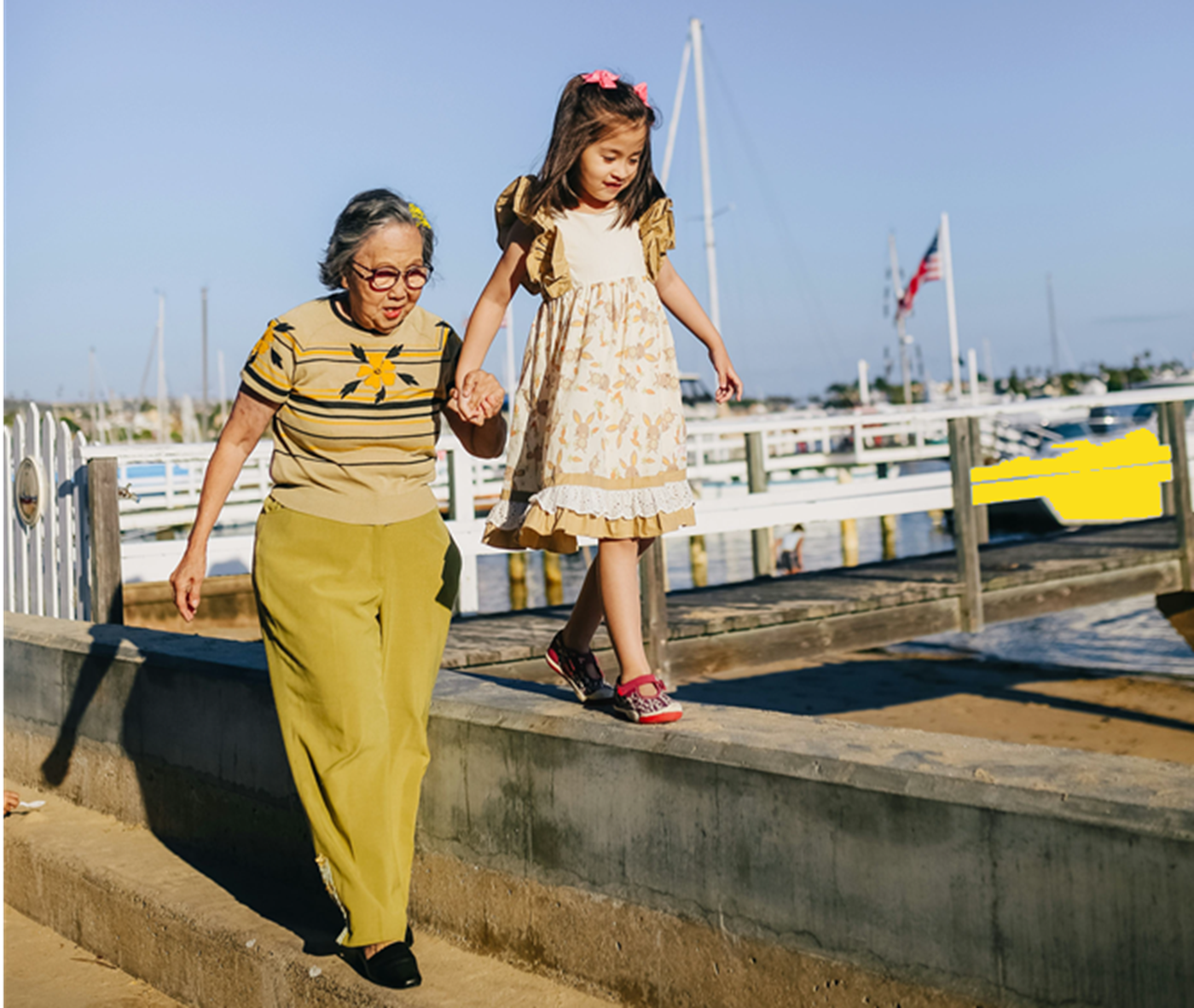 Elderly woman and young girl walking hand-in-hand on a cement ledge near a marina.