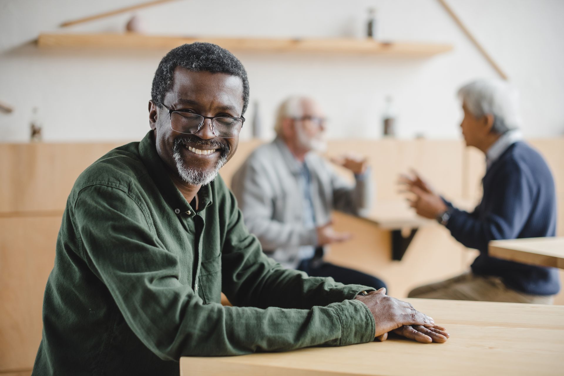 Smiling Black man at a table, two men conversing blurred in background.
