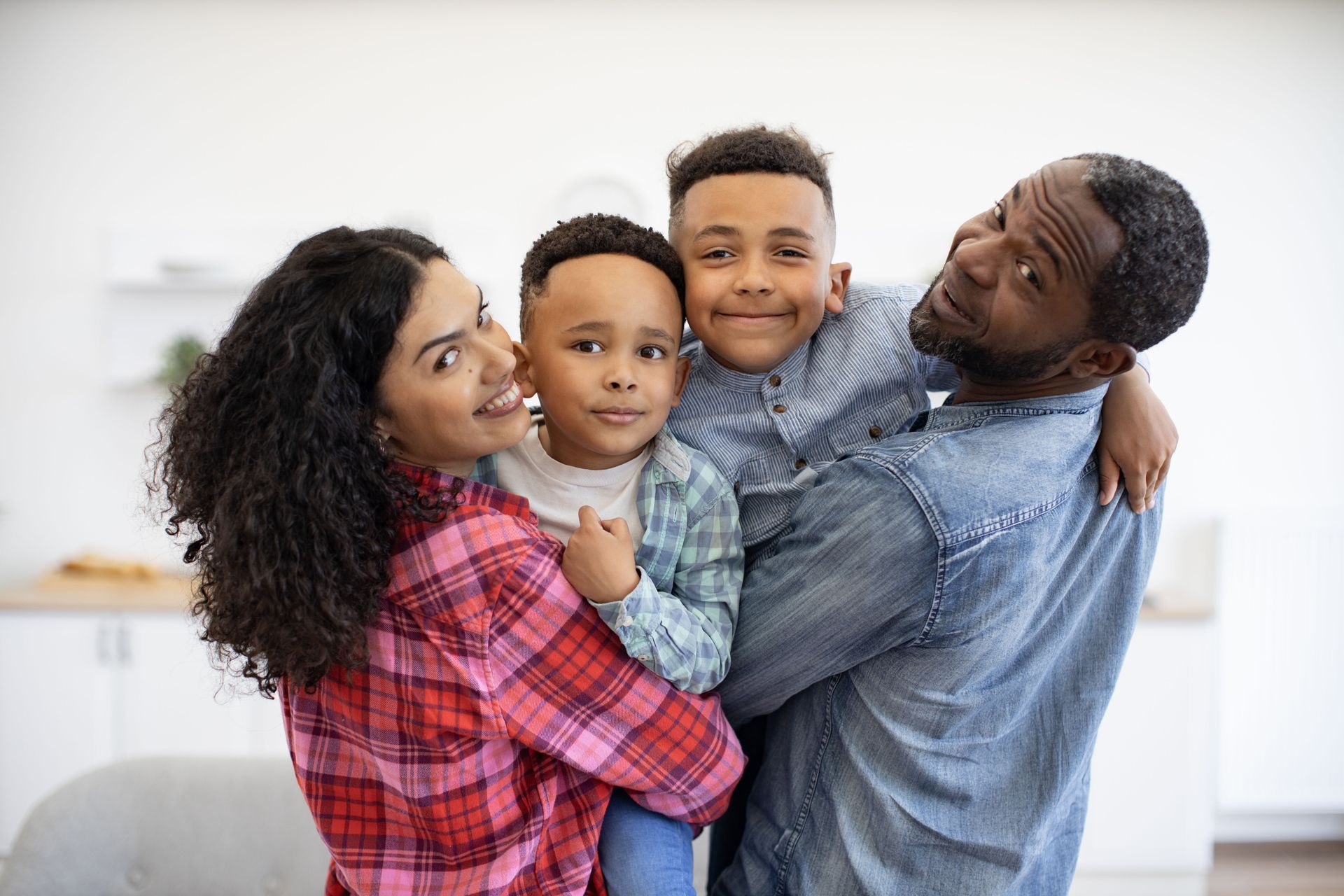 Family of four smiles: mom, dad holding two sons, indoors.