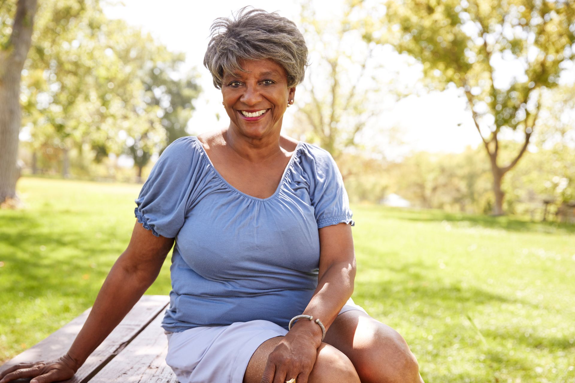 Smiling Black woman in blue shirt and white skirt sits on a picnic table in a park.