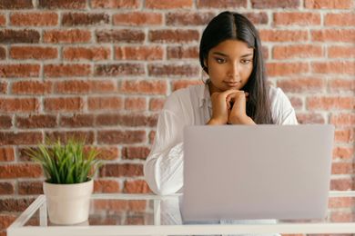 Woman at laptop with hands clasped, leaning on desk. Brick wall background, potted plant.