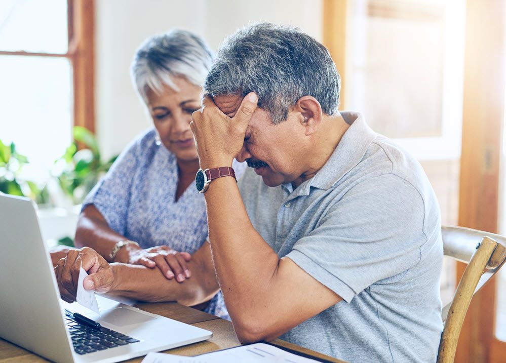 Older couple looks stressed at a laptop, woman comforting the man, both indoors.