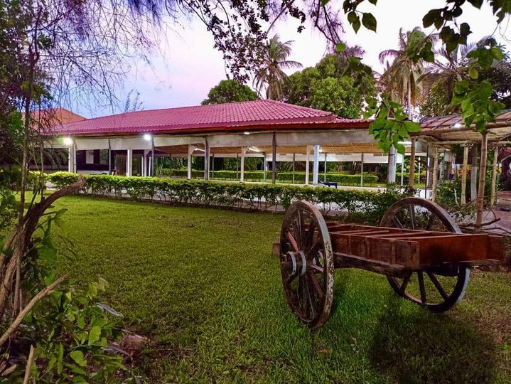 Un carro rústico de madera reposa sobre el césped frente a un edificio largo y bajo con techo rojo.