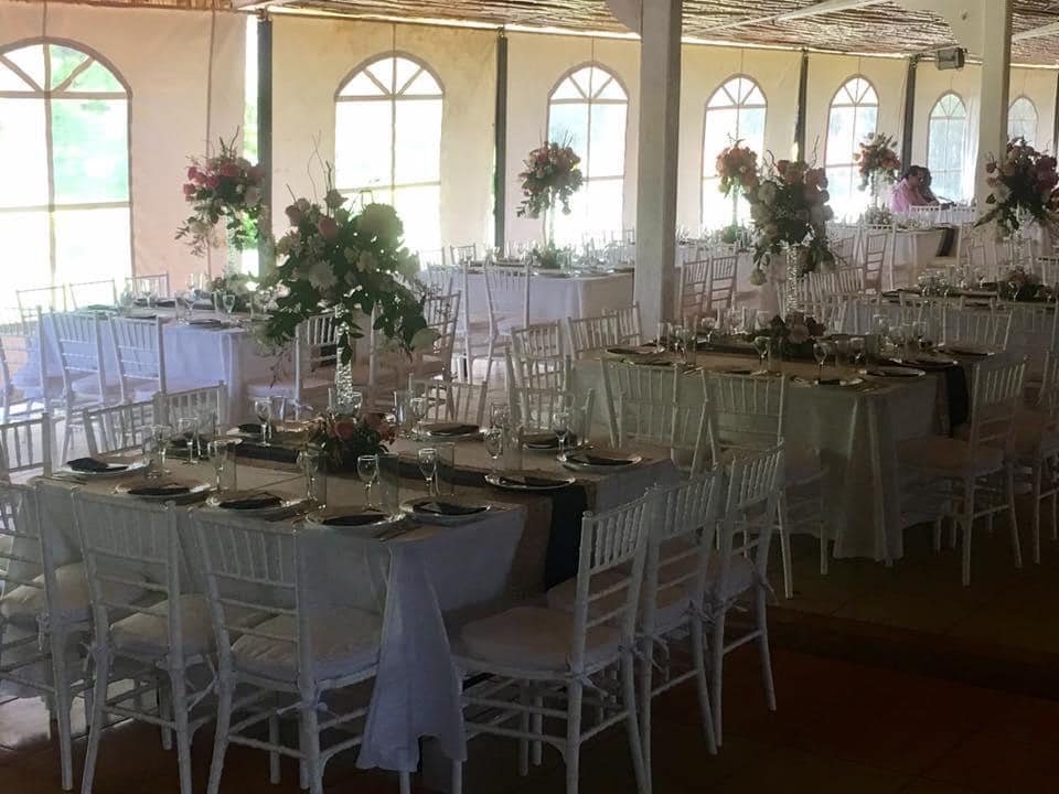 Salón de banquetes preparado para una celebración, con mesas dispuestas con mantelería blanca, flores y sillas blancas.